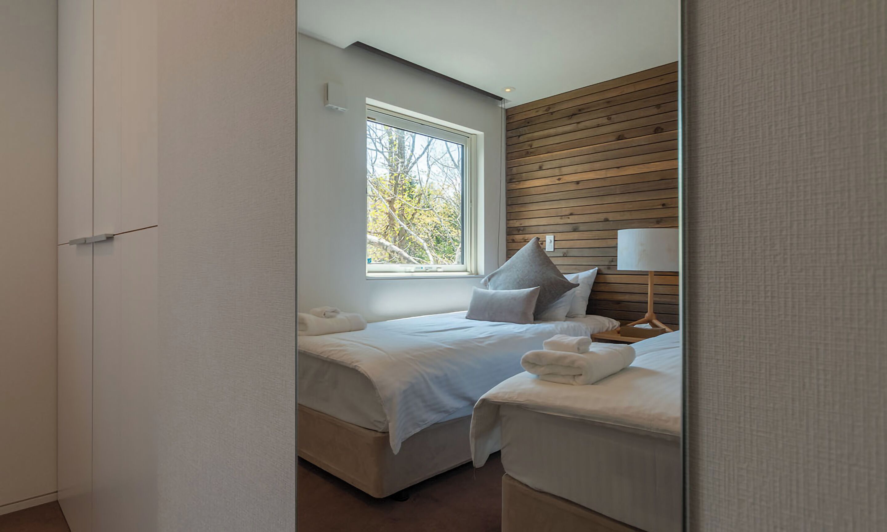 A bedroom with a large window, wooden panel wall, and a bedside table with a lamp.
