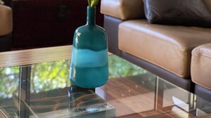 Ein Zimmer mit einem Glastisch, einer türkisfarbenen Vase mit Blumen, einem braunen Sofa und Blick auf Grünflächen.