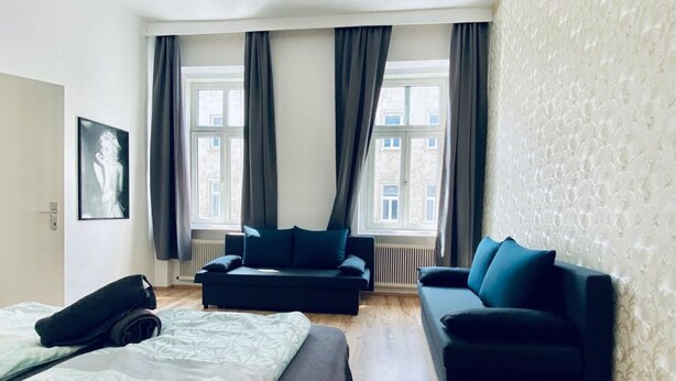 Habitació d'hotel amb un llit, dos sofàs, un radiador i grans finestres amb cortines.