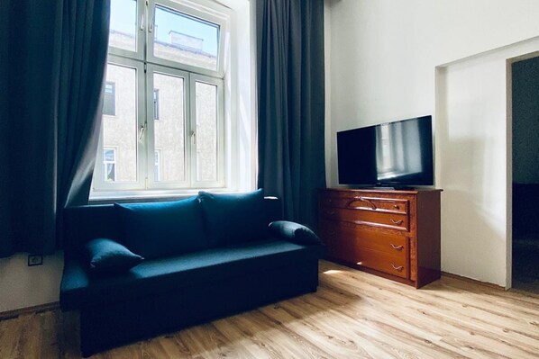 A room with a sofa, a TV on a wooden dresser, a window with curtains, and a view of buildings outside. - Comfy 3BR Apt In Prime Location (Vienna)