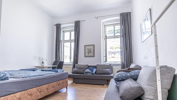Quarto com cama, sofá, duas janelas com cortinas, radiador e um quadro na parede.