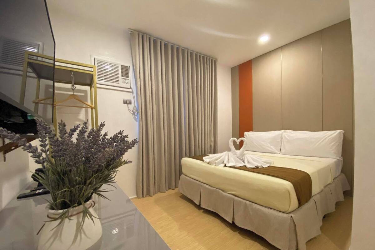 A modern hotel room with a bed, a potted plant, a mirror, and a window with curtains.