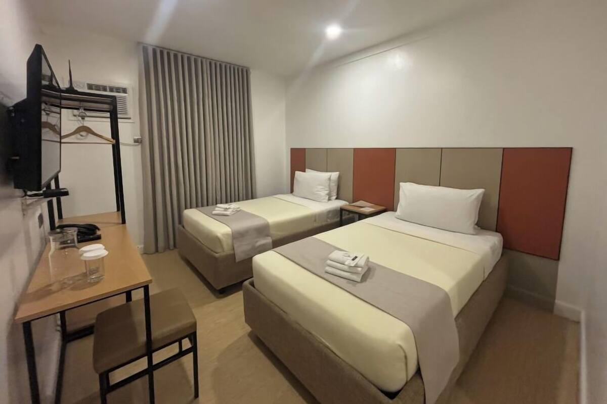 A hotel room with two beds, a desk, a chair, a TV, and a window with curtains.