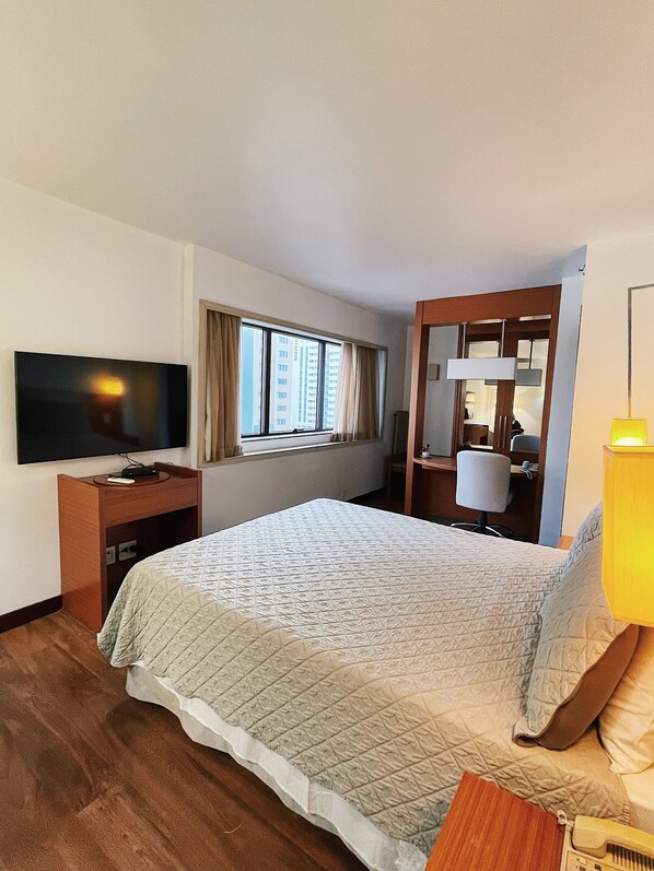 A hotel room with a bed, a TV on a wooden stand, a window with curtains, a desk with a lamp, and a mirror. - Quarto Hotel Comfort Suites Brasilia- o Mais Confortável e Aconchegante (Asa Norte)