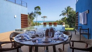 A round dining table set for a meal with a view of the ocean, surrounded by wicker chairs, a blue building, and a pool area.