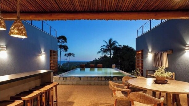 An outdoor dining area with a pool, wooden furniture, and a view of the ocean.