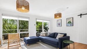 A modern living room with a blue sofa, a wooden chair, and a small table. There are large windows with a view of greenery outside.