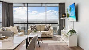 A modern living room with a sofa, coffee table, and TV. It features large windows with a view of the city and a beach. - Circle on Cavill River View (Surfers Paradise)