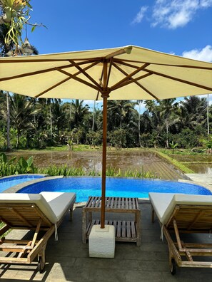 A poolside area with a wooden umbrella, lounge chairs, and a view of a lush garden and water body.