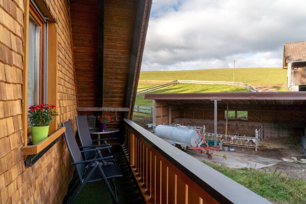 A balcony with a view of a farm, a wooden building, and a fenced area with farm equipment.