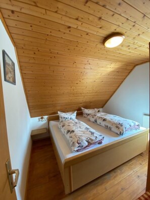 A small, single-bed room with wooden walls and ceiling, a white bed with patterned bedding, and a simple door.