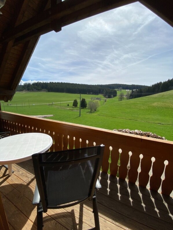 A wooden deck with a table and chair, overlooking a green field and forested hills.