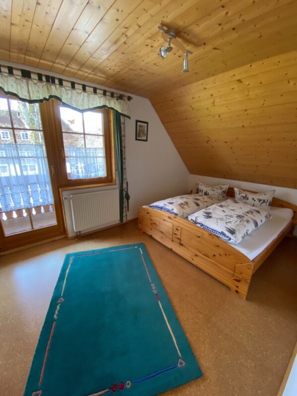 A bedroom with a wooden ceiling, a bed with patterned bedding, a window with curtains, and a blue rug. - Apartment 'Hofblick' with Shared Garden and Balcony, Farm Holiday (Eisenbach)