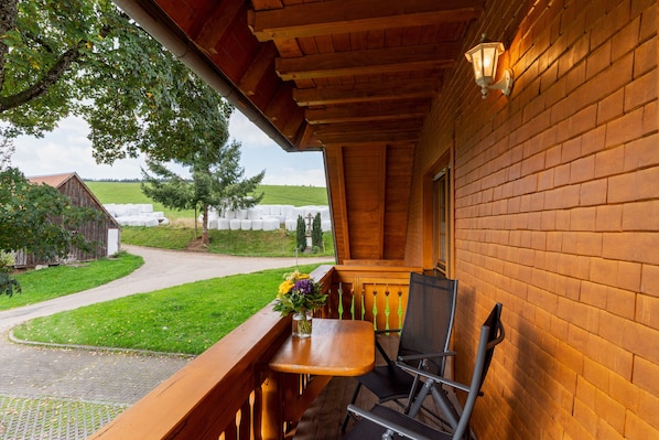 A wooden balcony with a table and chairs, overlooking a green landscape and a building with hay bales.
