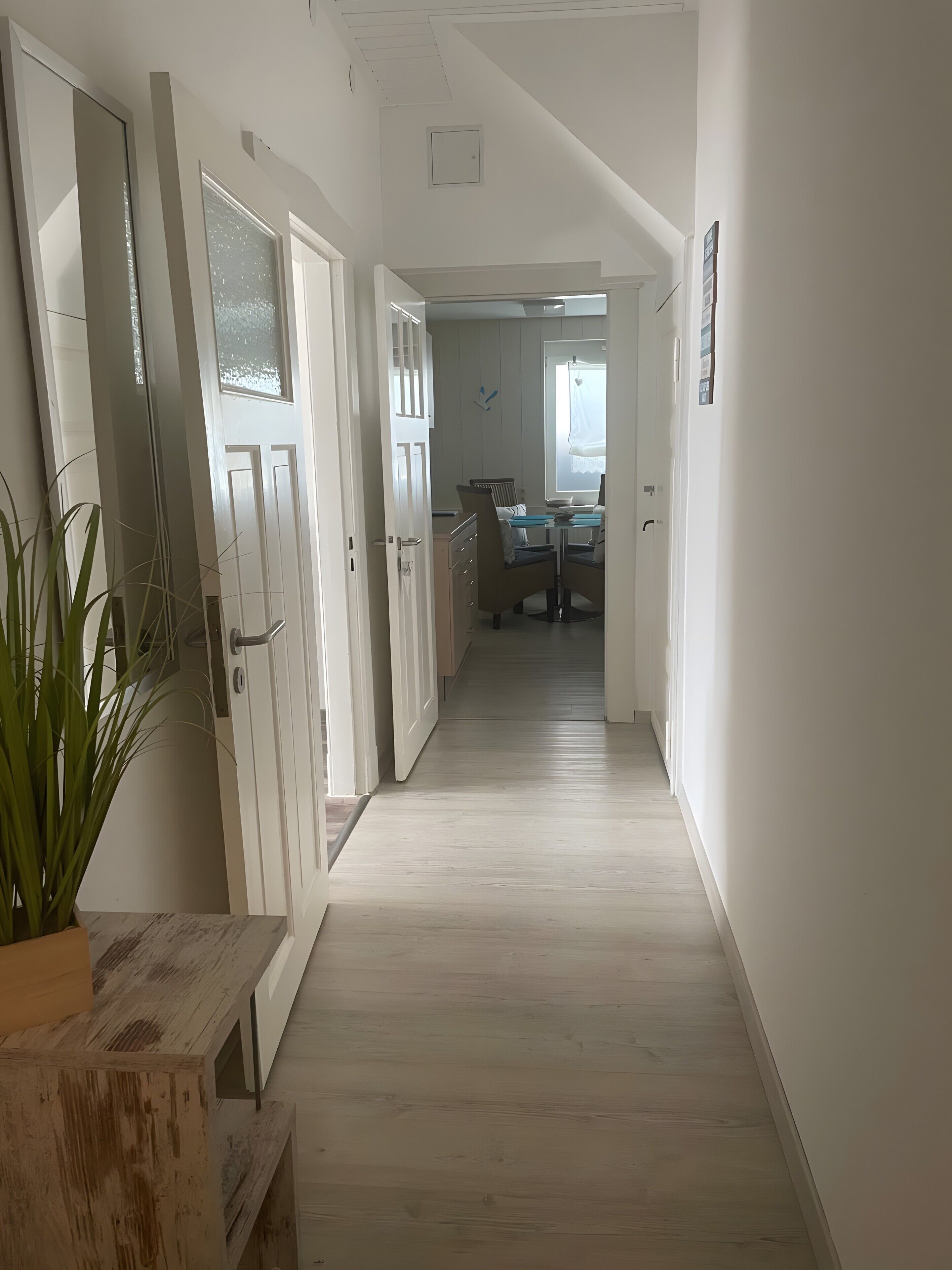 A hallway with a wooden floor, white walls, and a door leading to a dining area with a table and chairs.