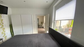Ein Schlafzimmer mit Bett, Kleiderschrank, Fenster mit Jalousien und Blick nach draußen.