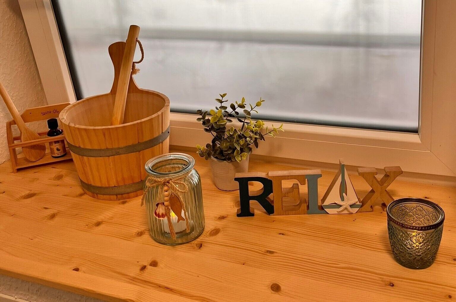 Een houten plank met een houten emmer, een glazen pot met een touwtje, een bloempot, houten letters die 'RELAX' vormen, en een kaars.