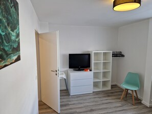 Une pièce avec un téléviseur posé sur un meuble blanc, un fauteuil de couleur turquoise et un plancher en bois.