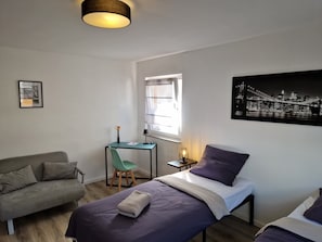 Une pièce comprenant un lit, un bureau, une chaise, un canapé, une fenêtre et un tableau représentant un paysage urbain accroché au mur.