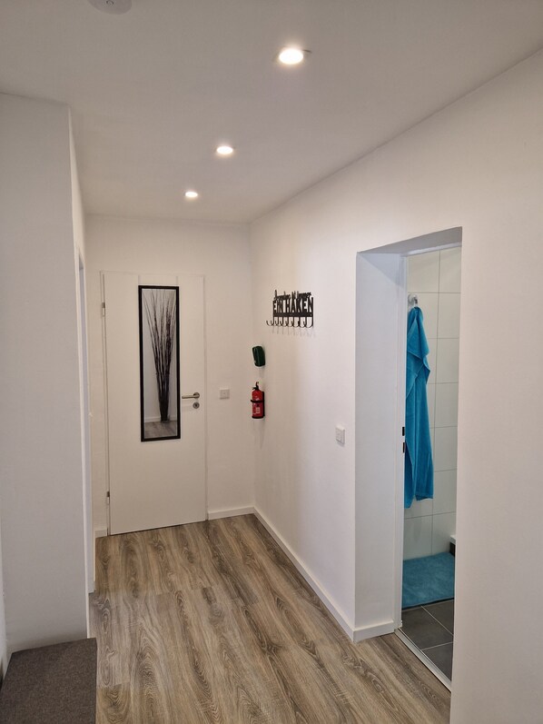 A hallway with a wooden floor, a white door, a framed picture, a wall-mounted fire extinguisher, and a towel hanging on the wall.
