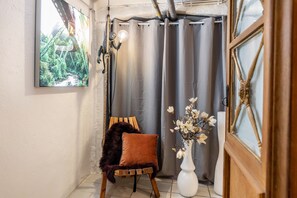 Une pièce avec vue sur la verdure, un fauteuil recouvert d’un plaid en fourrure, un vase blanc orné de fleurs et une porte en bois.