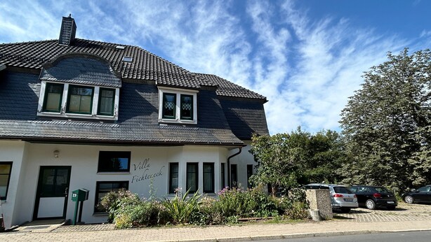 A two-story house with a dark roof, white walls, and a small garden with flowers. - Studio Apartment 'Wohnung 1' with Private Terrace and Wi-Fi (Oberhof)
