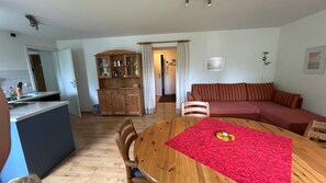 Ein Wohnzimmer mit Esstisch aus Holz, einer Couch und einer Küchenzeile mit Schränken und einem Kühlschrank.