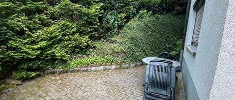 A paved courtyard with a metal chair and a table, surrounded by dense greenery and a building wall.
