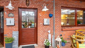 Un edificio de ladrillo con puerta de madera, dos lámparas fijadas a la pared, una maceta azul y una silla de mimbre en el exterior.