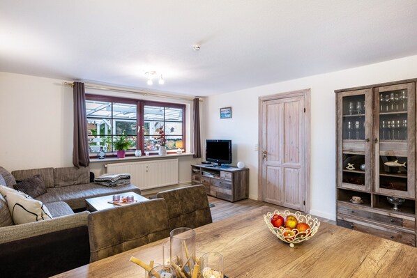 Ein Wohnzimmer mit Sofa, einem Esstisch mit Obstschale, einem Fernseher und einer Vitrine mit Gläsern.