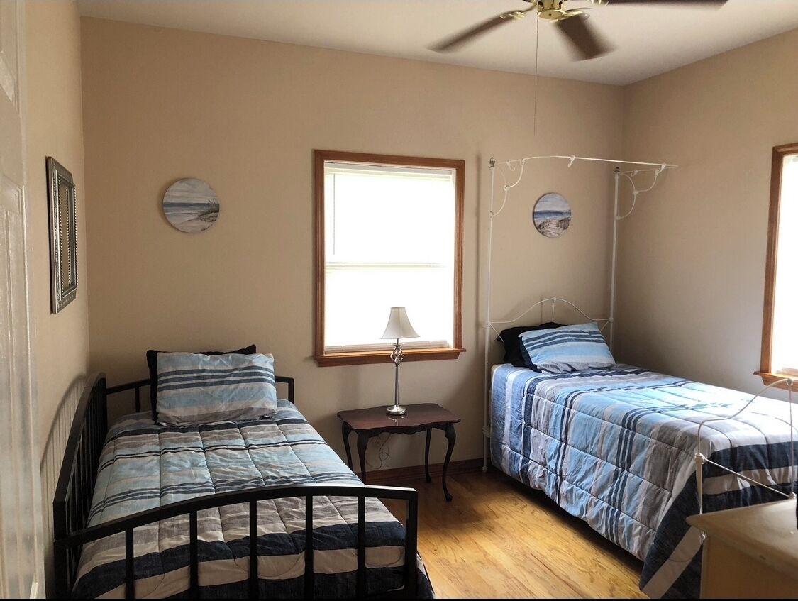 A bedroom with two single beds, a ceiling fan, a window with blinds, and a small table with a lamp.
