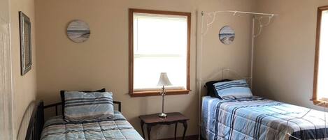 A bedroom with two single beds, a ceiling fan, a window with blinds, and a small table with a lamp.