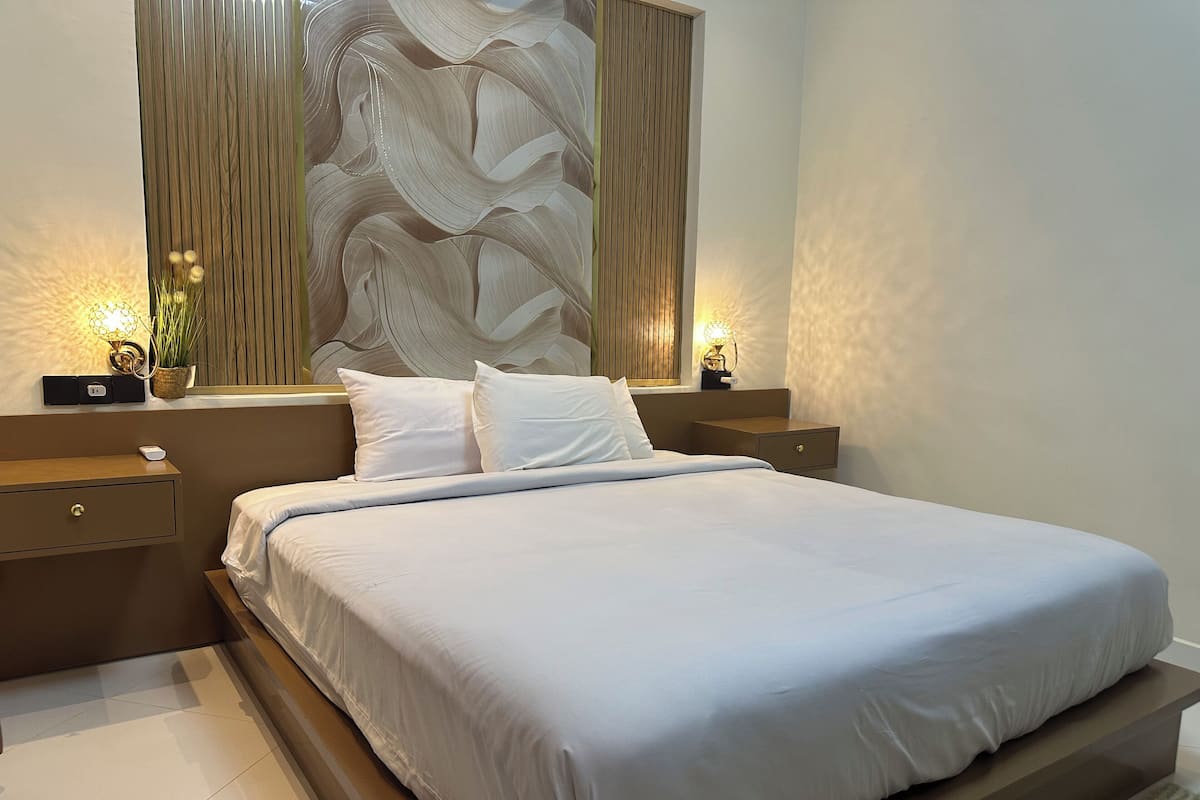 A modern bedroom with a large bed, wooden headboard, bedside tables with lamps, and a decorative wall panel.