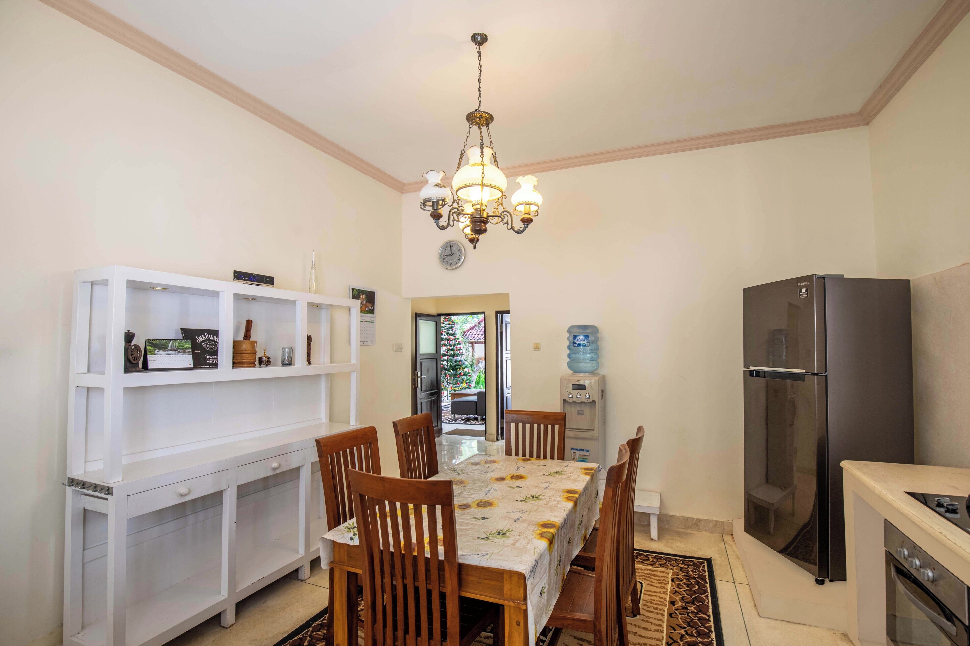 A dining area with a table set for six, surrounded by wooden chairs. A white console table with shelves is to the left, and a black refrigerator is to the right.