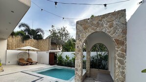 A pool area with a stone archway, lounge chairs, and a bamboo fence. - Ombe Villa (Aikmel)