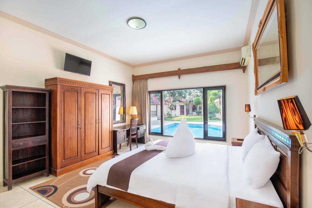 A hotel room with a large bed, a desk, a wardrobe, a TV, and a view of a swimming pool.