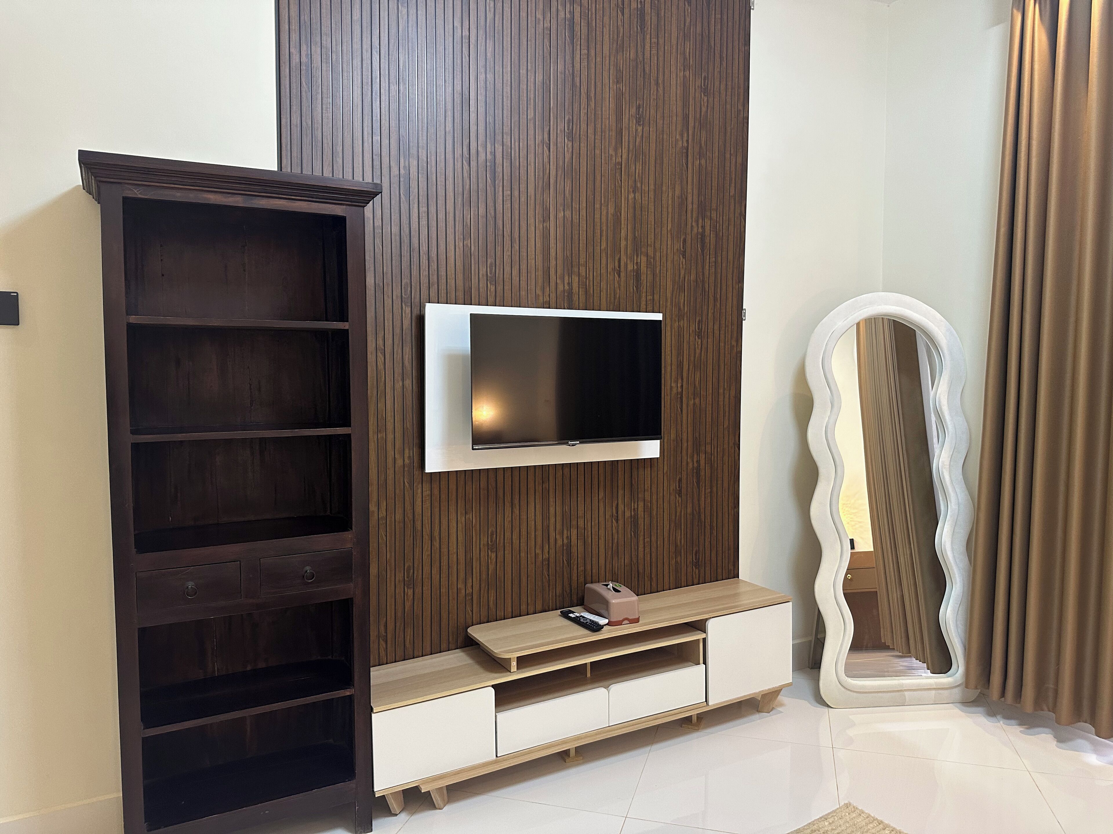 A modern living room with a TV mounted on a wooden panel wall, a large mirror, and a dark wooden cabinet.