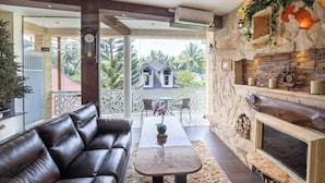 A modern living room with a leather sofa, a coffee table, and a fireplace. There is a balcony with a view of palm trees and a house. - Ombe Villa (Aikmel)