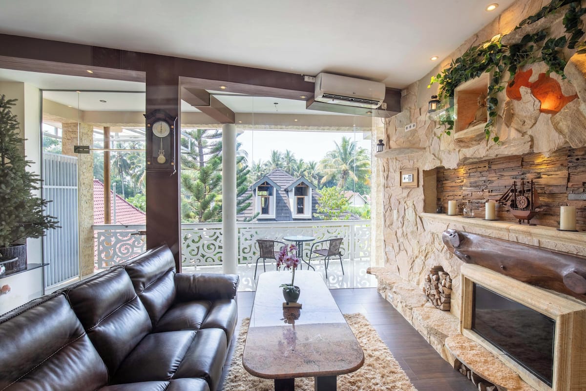 A modern living room with a leather sofa, a coffee table, and a fireplace. There is a balcony with a view of palm trees and a house.