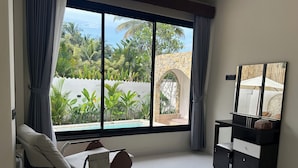 A modern bedroom with a large window, a rocking chair, a bedside table, and a built-in wardrobe. - Ombe Villa (Aikmel)