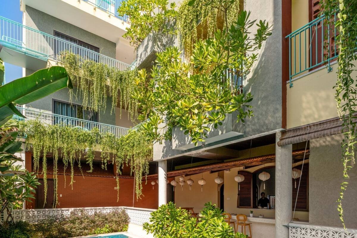 A swimming pool with a tiled deck, surrounded by greenery and a building with balconies.