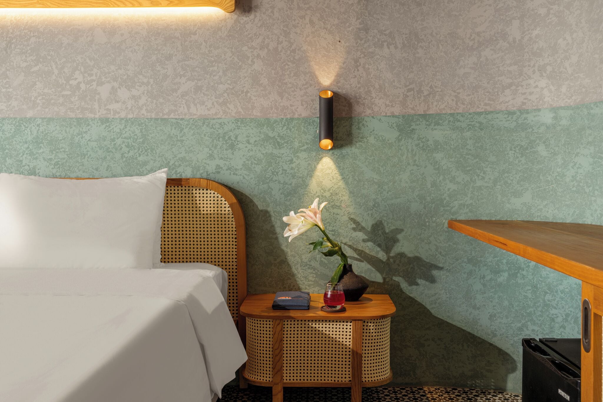 A hotel room with a bed, a small round table with a vase of flowers, a lamp, and a wooden side table.