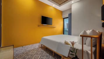 A hotel room with a yellow wall, a bed with white bedding, a mounted TV, a small table with a vase and candle, and a patterned floor.