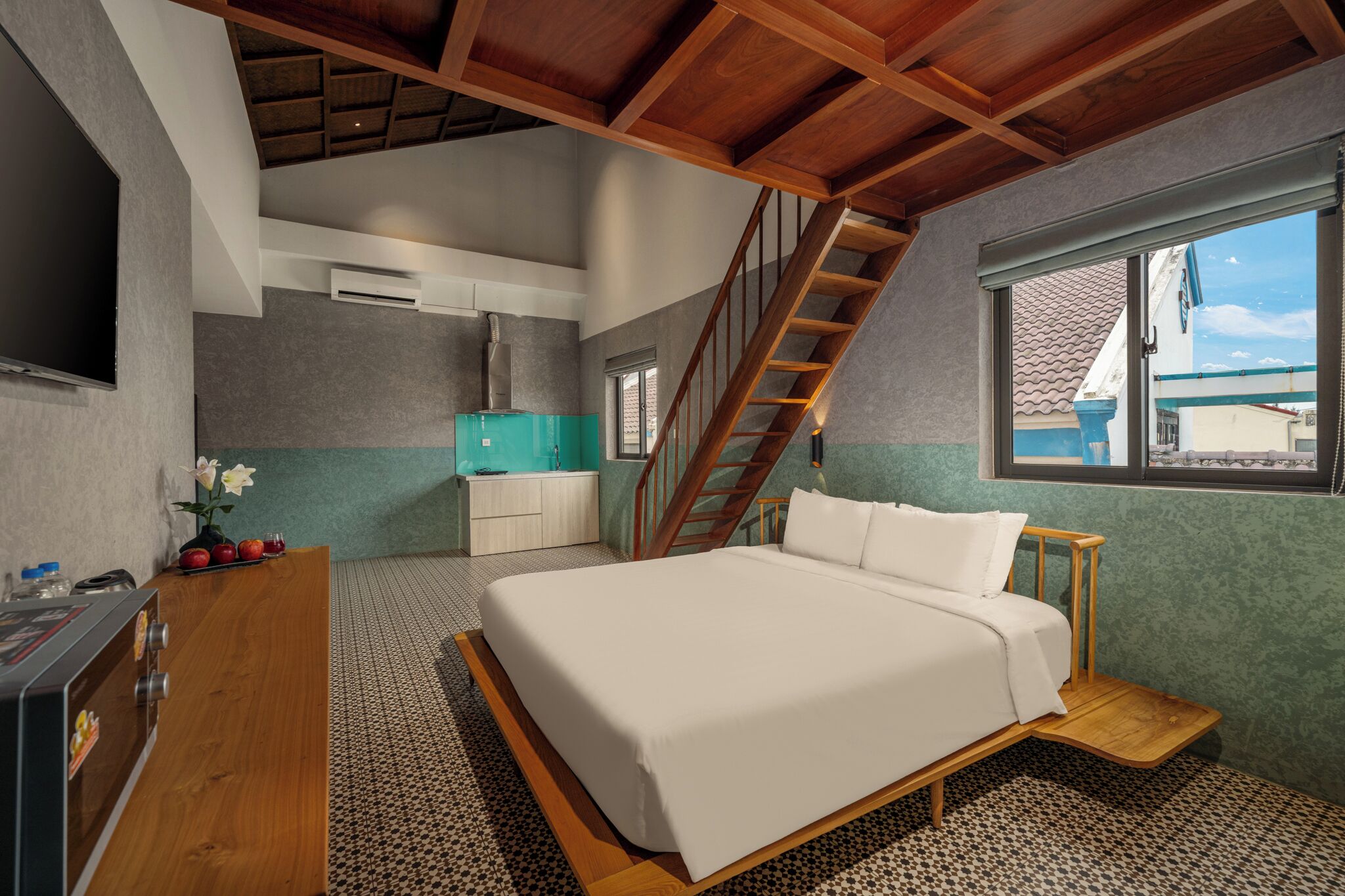 A bedroom with a bed, a wooden desk, a small kitchenette, and a staircase.