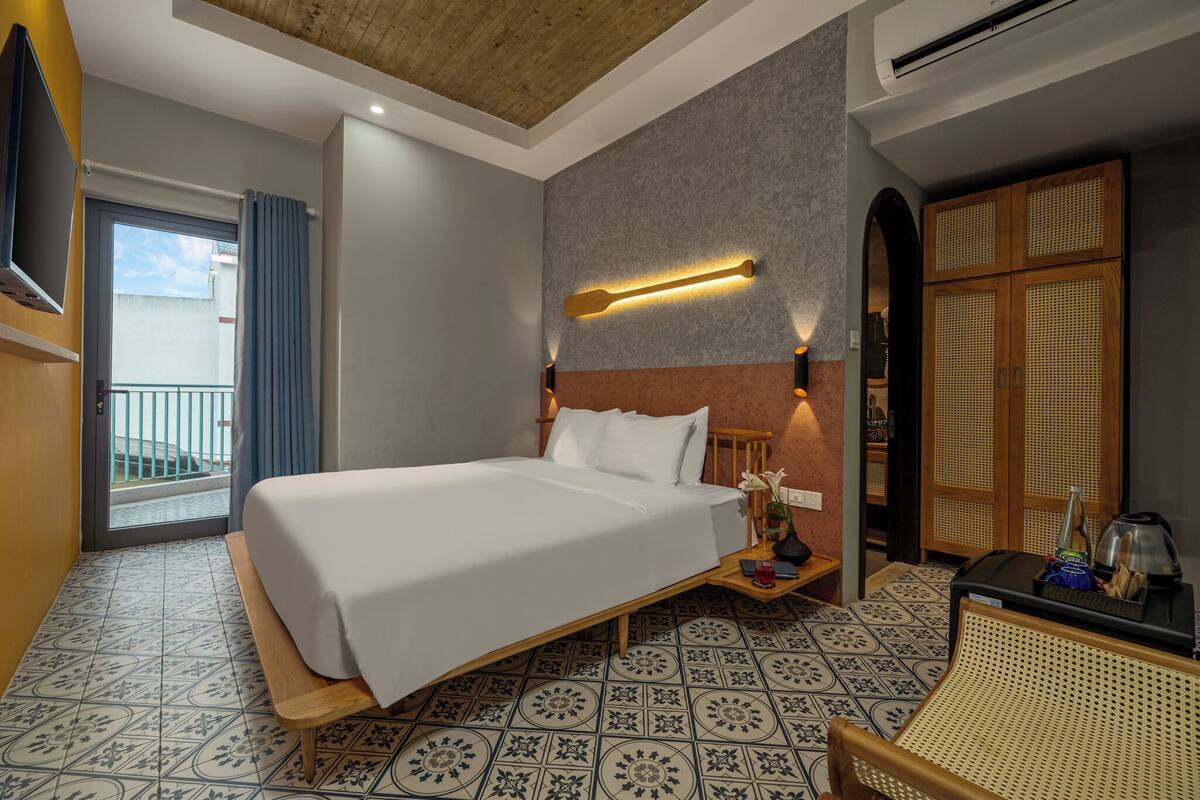 A hotel room with a bed, a nightstand, a wicker chair, a TV, and a balcony.