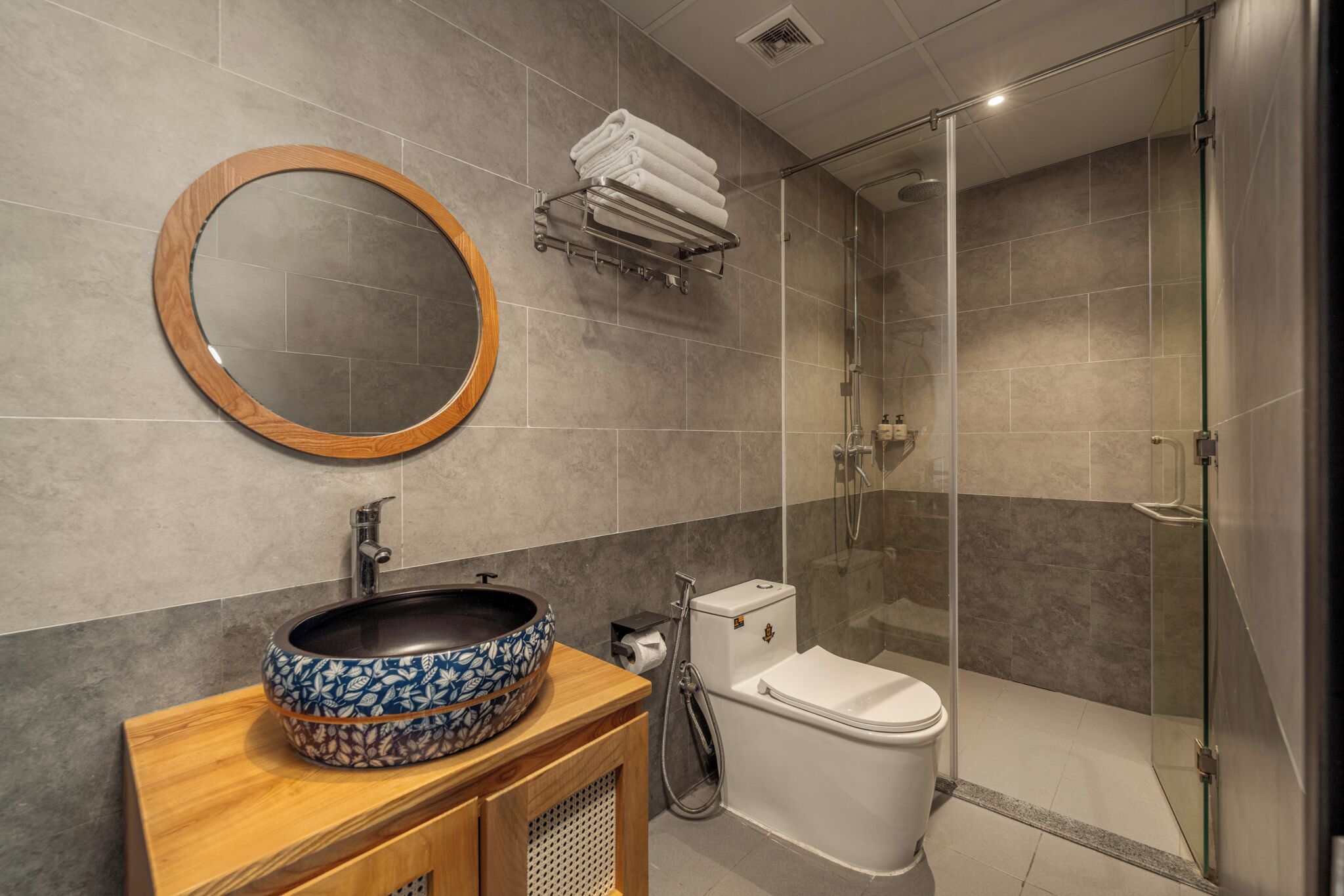 Une salle de bain moderne avec un grand miroir rond, un meuble-lavabo en bois, un grand lavabo décoratif en forme de bol, des toilettes et une douche à parois de verre.