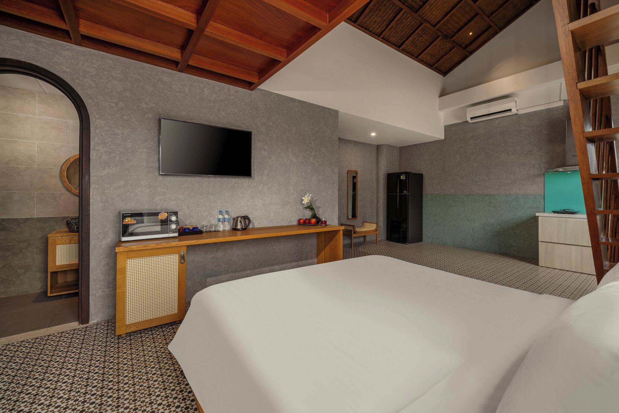 A modern hotel room with a large bed, a flat-screen TV mounted on the wall, a wooden desk with a mirror, a small kitchenette, and a wardrobe.
