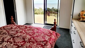 Un dormitorio con una cama grande, un aparador de madera y vistas a una playa a través de un ventanal corredero.