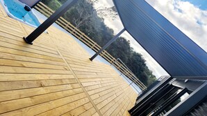 Una terraza de madera con piscina, barandas de metal y vistas a árboles y colinas.
