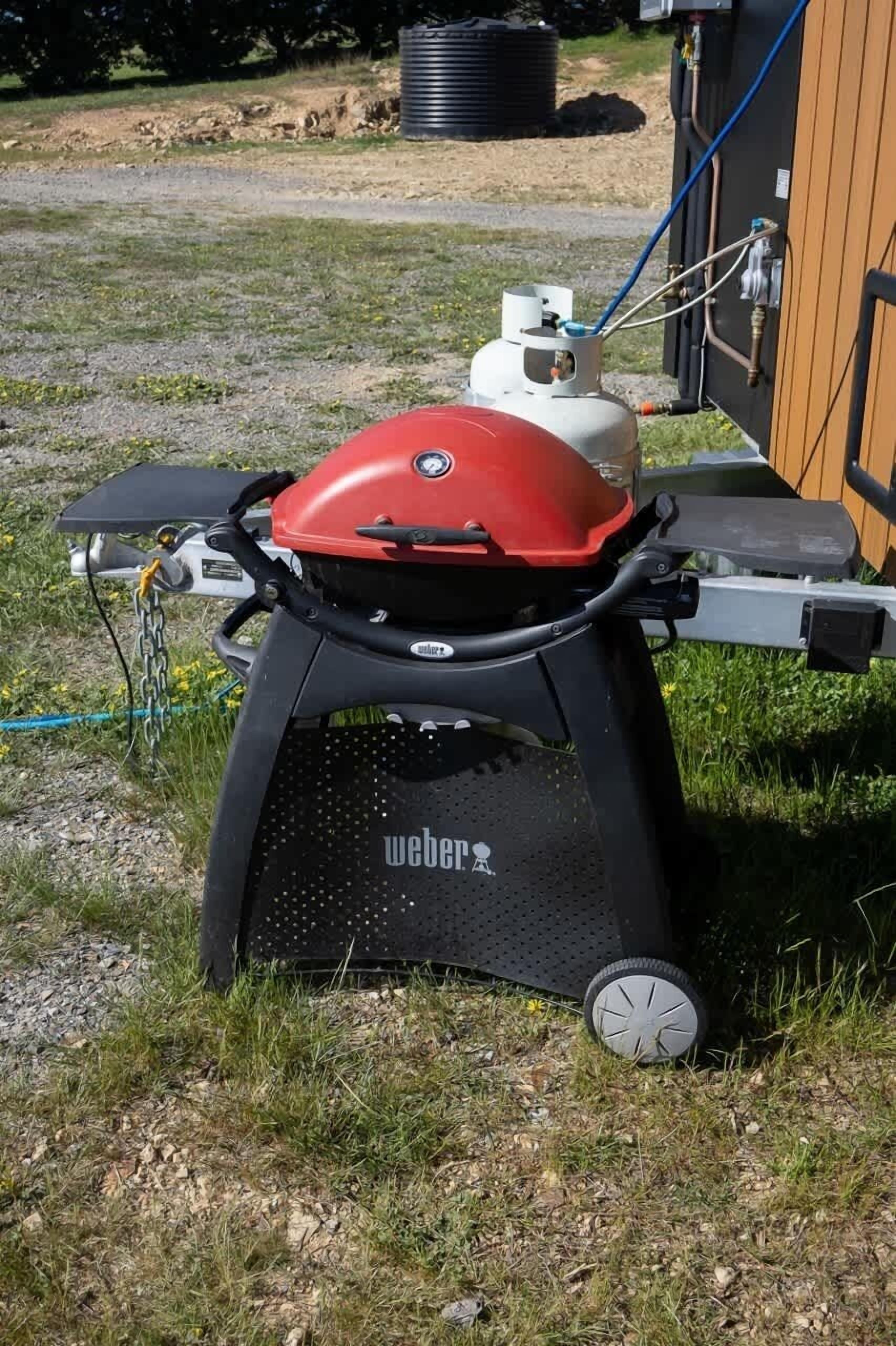 Una parrilla portátil con parte superior roja, base negra y ruedas, ubicada sobre césped cerca de una estructura de madera.